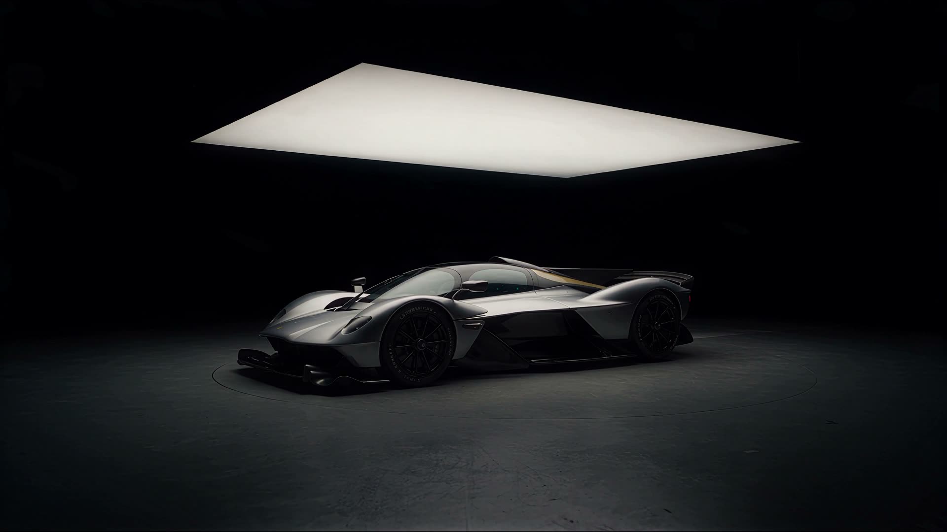 Silver hypercar standing in the middle of a black studio, lit by a single large suspended light panel.