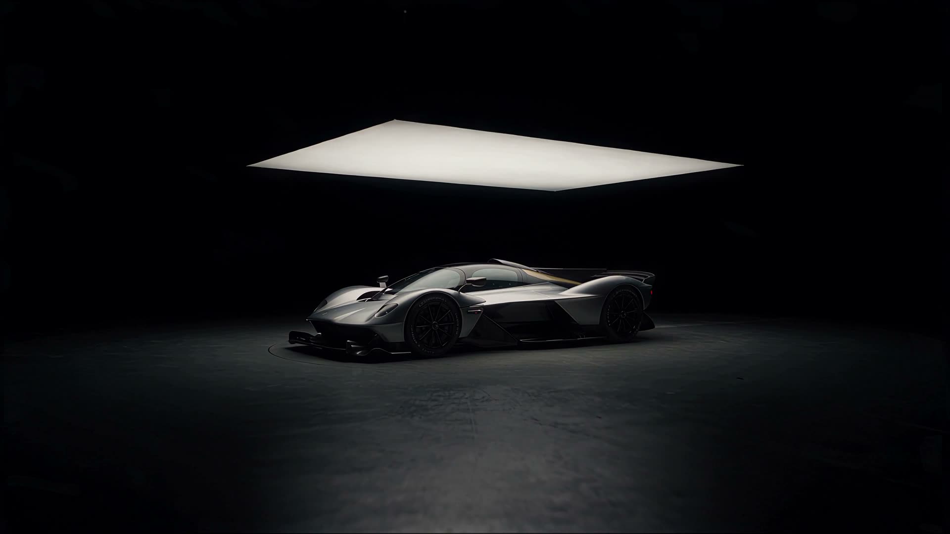Silver hypercar in three-quarter view in a black studio, under a large light panel.